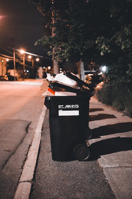 A row of five black wheeled rubbish bins with yellow lids, positioned on a concrete surface against a plain white wall. Each bin has a white oval label with identifying numbers, and the bins are evenl